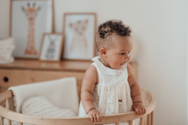 混合种族蹒跚学步的女孩在圆形木制婴儿床的肖像在舒适的儿童室中性色调图片下载
