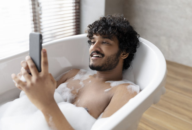 放松的印度男子躺在有泡沫的浴缸里，一边用智能手机，一边在温暖的泡沫水中放松图片下载