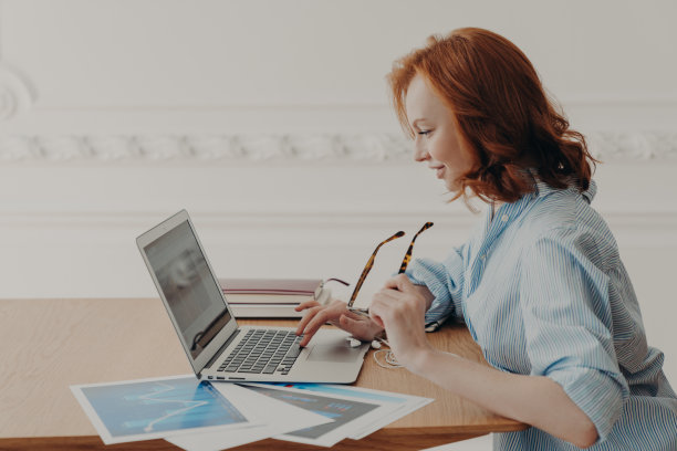 侧身镜头:红头发的年轻白人自由职业者远程工作，专注于笔记本电脑，坐在放着文件的木桌前，摘下眼镜，上网，看新闻图片下载