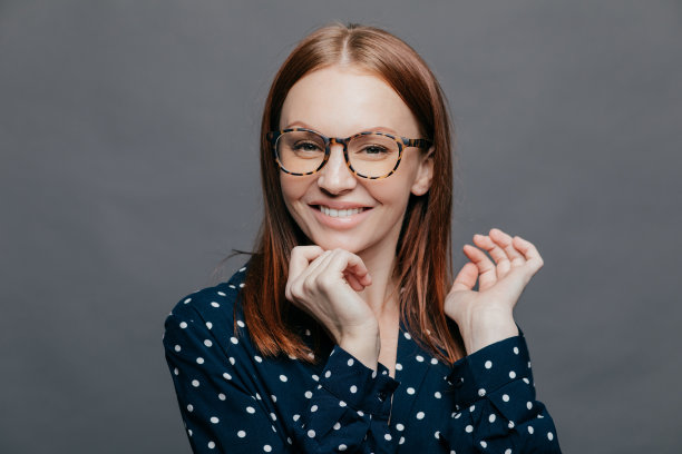 积极有吸引力的白人女子保持手在下巴附近，积极地看着相机，手在下巴下，笑得很开心，穿着白色圆点的黑色衬衫，在灰色背景上摆姿势。图片下载