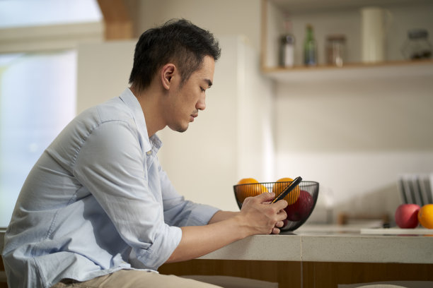 年轻的亚洲男子坐在厨房柜台看手机图片下载