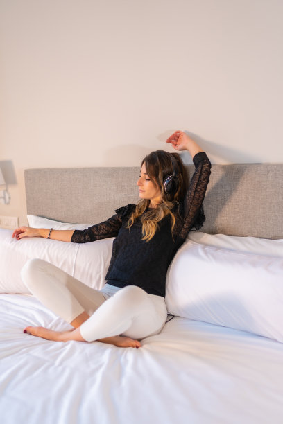 一个年轻女子坐在床上，戴着耳机享受音乐的垂直镜头图片下载
