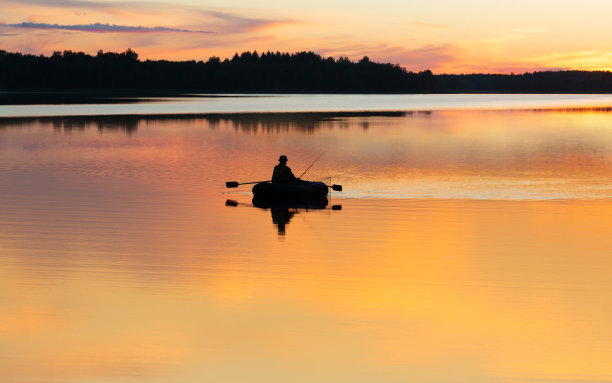 男人在日落或日出的时候从船上在湖上钓鱼。夕阳下渔夫的剪影图片下载