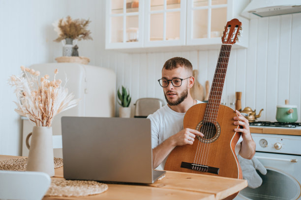 年轻的意大利大胡子，戴眼镜，坐在桌子前，拿着笔记本电脑，把吉他点在弦上，在家里，厨房上吉他课。爱好，远教育。远程学习。图片下载