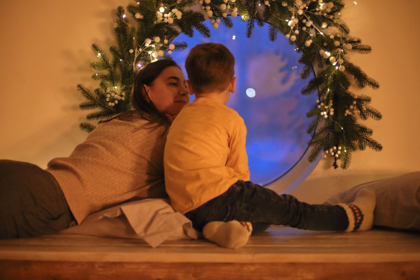 家庭母亲和小儿子在圣诞节前夕在家里看着窗外的后视图，选择性的焦点。圣诞晚上，妈妈和孩子坐在窗台上，老旧的灯笼闪闪发光图片下载