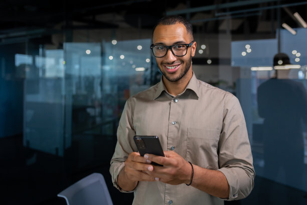 肖像年轻的西班牙裔男子在办公室里，一名穿着休闲衬衫的男子微笑着看着镜头，一名商人手里拿着智能手机，在电话上打字，打电话图片下载