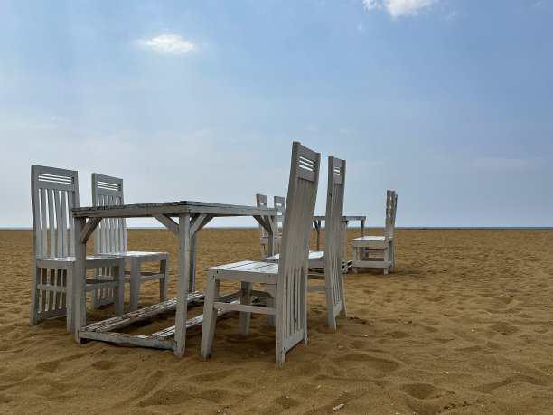 斯里兰卡首都科伦坡拉维尼亚山海滩上的露天用餐区，金色沙滩上质朴的木质板条餐桌特写，一组漆成白色的木制桌椅，蓝天白云背景，复制空间图片下载