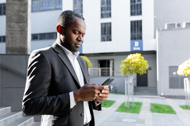 认真周到的商人在手机上阅读在线新闻，西装革履的非洲裔美国老板在办公大楼外盯着智能手机屏幕，在背景中走在大楼附近图片下载