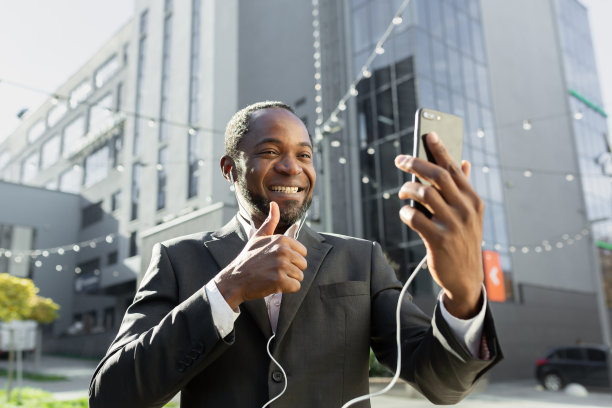 身穿西装、戴着耳机的非洲裔商人站在办公中心外，正在进行视频通话。手里拿着电话。对着镜头微笑，表现得超级棒图片下载