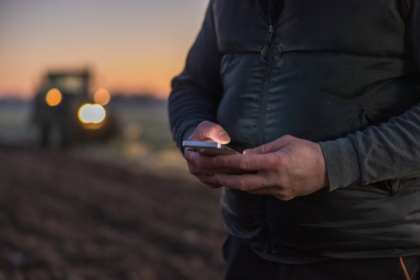 特写男性农民站在他的农田上使用智能手机，没有可见的脸，背景是拖拉机，车上的灯开着图片下载