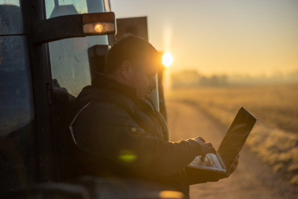 傍晚，一名棕色短发男子正靠在拖拉机上使用笔记本电脑，阳光从背后照进来，是他的剪影图片下载