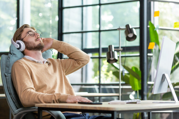 年轻的商人从工作中休息，加班，休息。坐在办公室的桌子前，对着电脑，靠在椅子上，戴着耳机听音乐，闭上眼睛，放松图片下载