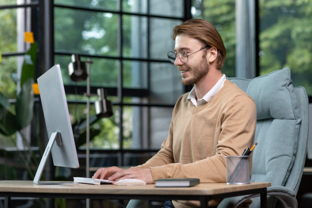 一位年轻的男性程序员，自由职业者，坐在办公室靠窗的桌子旁，用电脑打字。微笑着，戴着眼镜，穿着米色毛衣图片下载