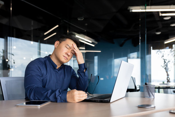 一个亚洲男人头痛得厉害。偏头痛。他坐在桌子旁，抱着头，闭上眼睛图片下载