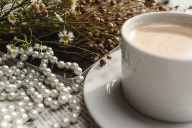 一杯白色的咖啡放在茶托上，上面有一串珍珠、干花、一本杂志和一份报纸。唯美的画面，优雅的背景照片。图片下载