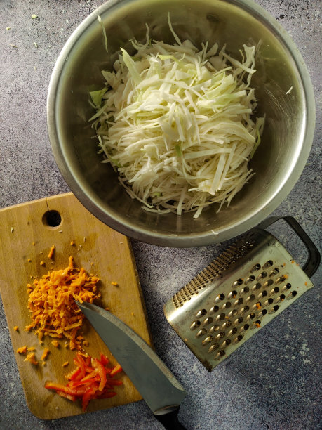 白色的卷心菜放在一个金属碗里，一个磨丝器和一个木板，上面有胡萝卜和青椒，放在灰色的表面上图片下载