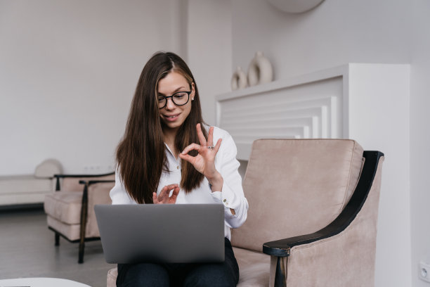 开朗的年轻黑发意大利女子笑着视频通话显示ok迹象，坐在客厅的椅子上。戴眼镜的西班牙漂亮女商人表示赞同。商业人士的概念。图片下载