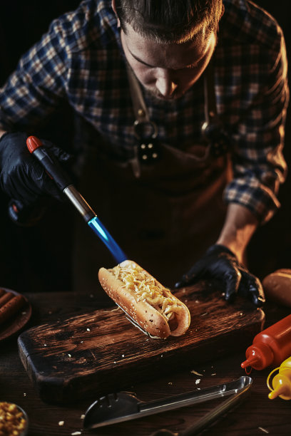 一个穿着围裙和手套的人在热狗上撒奶酪。做热狗。创造经典快餐。图片下载