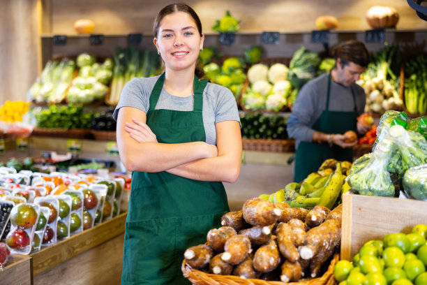 在超市水果和蔬菜的背景下，穿着围裙的友好女售货员的肖像图片下载