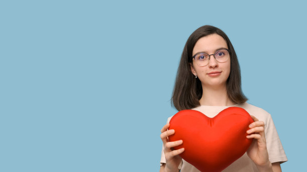 一个美丽的微笑的学生女孩的肖像，戴着眼镜，抱着一个鲜红色的心形枕头图片下载