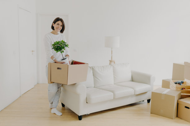 全程拍摄，开朗的黑发女子拿着有室内植物的纸箱，站在宽敞的房间里，有沙发和落地灯，穿着休闲装。女房客站在新家图片下载