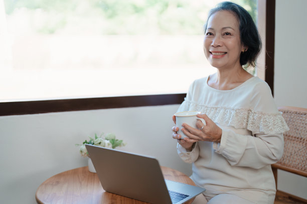 一个年长的亚洲妇女在现代姿态做电脑工作和喝咖啡的肖像。图片下载