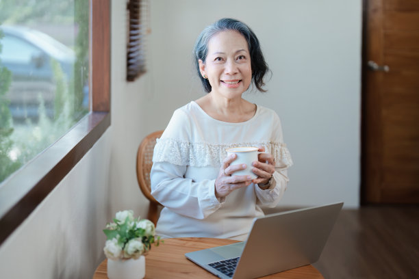 一个年长的亚洲妇女在现代姿态做电脑工作和喝咖啡的肖像。图片下载