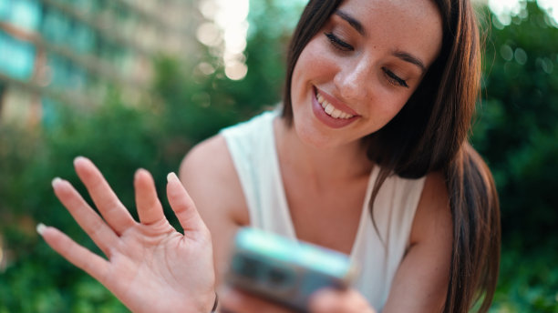 可爱的年轻女子雀斑和黑色松散的头发穿着白色上衣正在使用手机。美丽的女孩坐在城市公园看照片，视频在手机上。摄像机四处移动图片下载