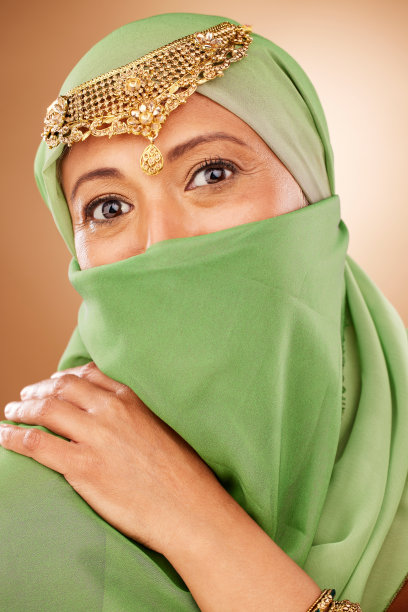 时尚、传统：一位戴着传统珠宝和罩袍的穆斯林女性在工作室里。美丽、伊斯兰：成熟女士戴着伊斯兰头巾或阿拉伯珠宝的肖像，棕色背景图片下载