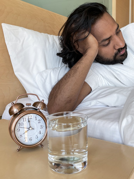 印度男人的形象靠在肘部在酒店双人床从羽绒被下由闹钟在床头柜，青铜金属案件，复古闹钟，双钟，模拟显示时钟，饮用水，重点在前景图片下载