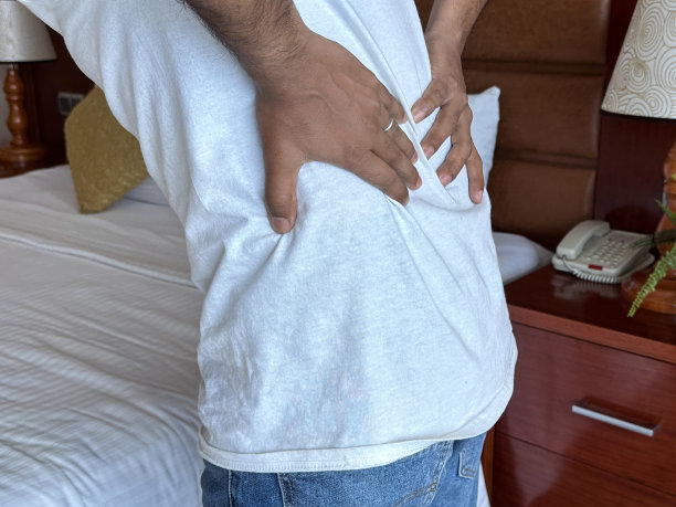 特写照片:一个不认识的人站在酒店双人床旁，双手放在腰上，痛苦地蜷缩在床上，腰酸背痛，有盆栽的床头柜，电话，灯聚焦在前景，假期和假期疾病的概念图片下载