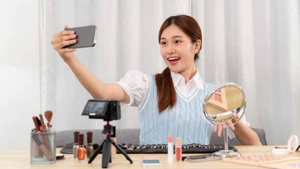 年轻的美妆网红一边用智能手机自拍，一边用美容设备和化妆配件图片下载