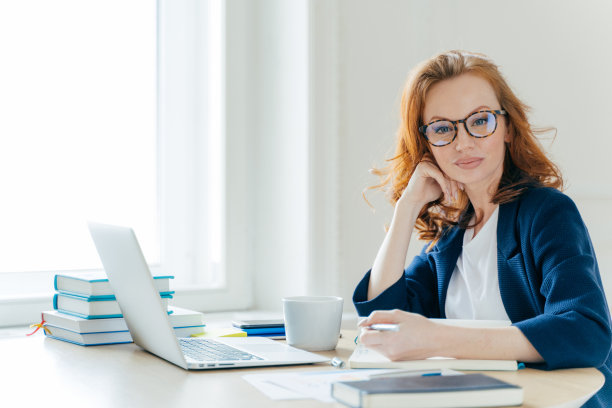 自信成功的红发女企业主坐在橱柜前，在笔记本电脑上工作，在笔记本上写主要论文，有咖啡休息，在宽敞的办公室里摆姿势图片下载