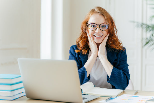 积极的红头发行政经理在互联网网站上搜索信息，触摸脸颊，在记事本上写笔记，使用现代笔记本电脑，穿着正式的衣服，检查组织者。图片下载