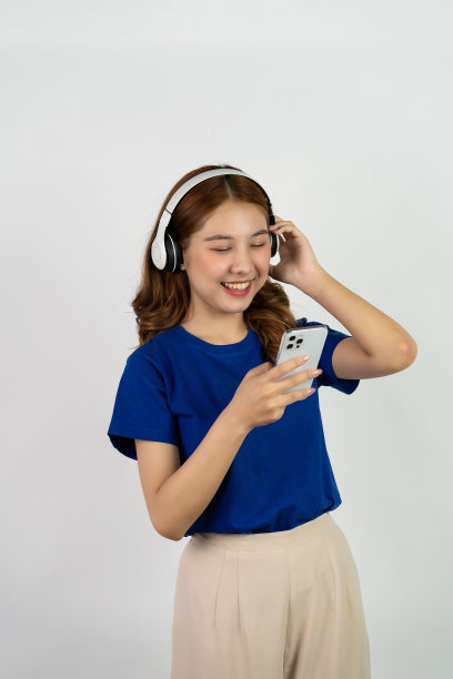 美丽的年轻女子拿着手机，戴着耳机，听着音乐，脸上带着微笑，白色背景。在线娱乐与音乐流媒体的概念图片下载