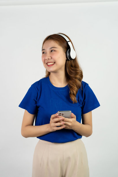 美丽的年轻女子拿着手机，戴着耳机，听着音乐，脸上带着微笑，白色背景。在线娱乐与音乐流媒体的概念图片下载