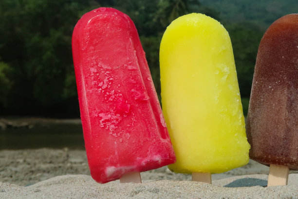 近距离图片，三排冰棒和木制冰棒棒卡在沙滩上，退潮时，红色，黄色和棕色的冰棒在海边的水边，草莓，柠檬和可乐口味的冰棒在海岸背景下，重点在前景图片下载