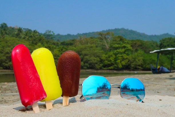 三排冰棒的特写图片:粘在沙滩上的木制冰棒，退潮时海边的红色、黄色和棕色冰棒，草莓、柠檬和可乐口味的冰棒，以海岸为背景，镜像太阳镜，复制空间图片下载