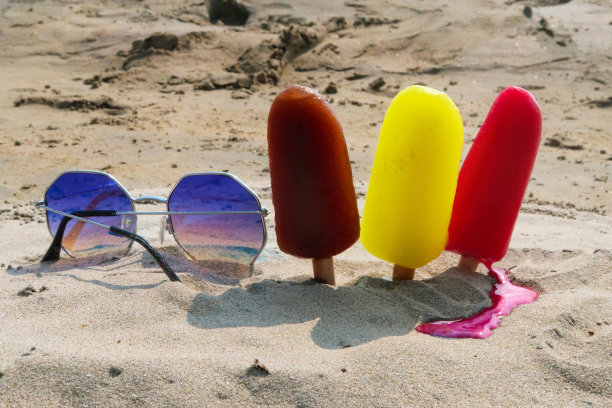 近距离图片，三排冰棒，木制的冰棒棒卡在沙滩上，红色，黄色和棕色的冰棒除了镜像太阳镜，融化的草莓，柠檬和可乐口味的冰棒，重点放在前景图片下载