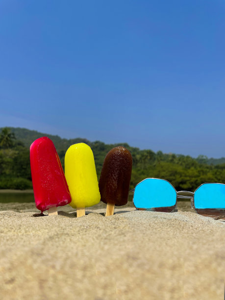 第三排冰棒的图片——粘在沙滩上的木制冰棒，退潮时海边的红色、黄色和棕色冰棒，草莓、柠檬和可乐口味的冰棒，以海岸为背景，镜像太阳镜，复制空间图片下载
