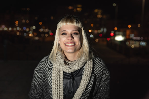 美丽的年轻时尚潮女孩的夜晚肖像与围巾和帽子在外套与城市路灯的背景图片下载