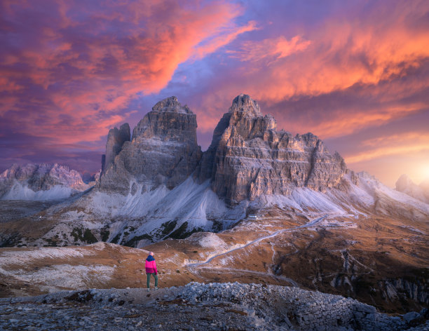 女人在山上和山峰在秋天五颜六色的日落。在意大利的白云石徒步旅行。小路上的女孩，秋天黄昏的高高的岩石。有悬崖峭壁的风景，紫色的天空和粉红色的云图片下载