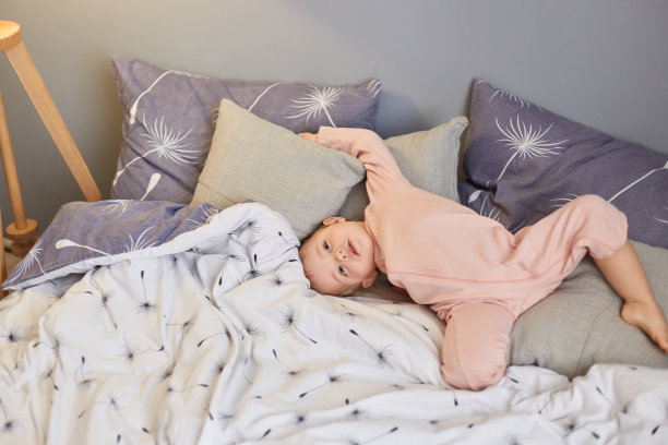 迷人的有趣可爱的婴儿女婴躺在卧室里，带着好奇的表情，在床上享受慵懒的早晨，在枕头上摆姿势。快乐的童年。图片下载