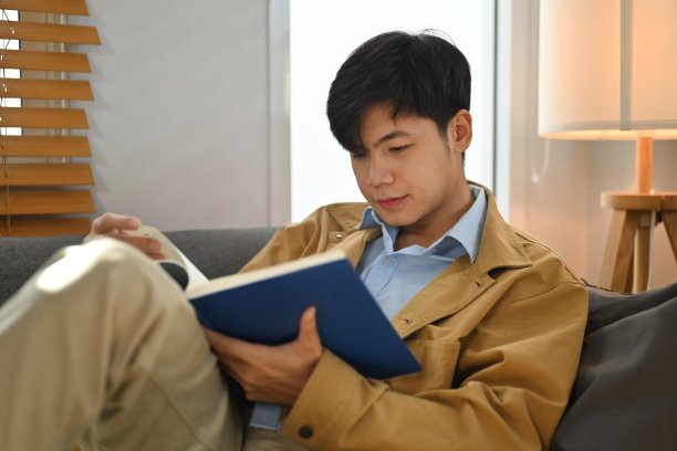 冷静的亚洲男人坐在舒适的沙发上，读着有趣的书，在家里享受悠闲的周末时光。图片下载