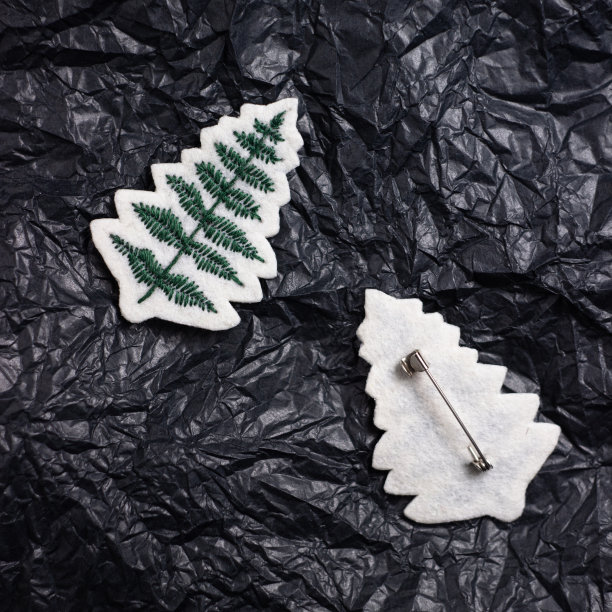 胸针绣有线在白色的感觉在一个蕨类的形式，旁边躺着它的背面与一枚针。背景是皱巴巴的黑纸图片下载