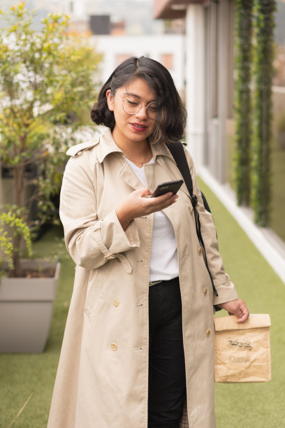 垂直镜头下，这位年轻的短发女子穿着奶油色外套，拿着手机和一个纸午餐袋。图片下载