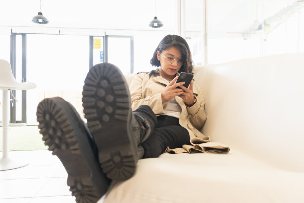 一个穿着奶油色外套的年轻短发女人坐在沙发上看手机。图片下载