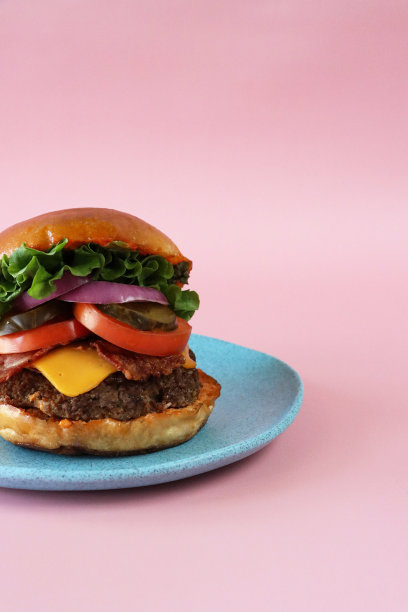 图片自制的培根芝士汉堡面包面包在绿松石盘，汉堡肉饼，培根片，融化加工奶酪片，生菜叶，切片番茄，泡菜和红洋葱片，粉红色背景，重点在前景，复制空间图片下载