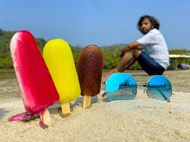 第三排冰棒的特写图片，木制的冰棒卡在沙滩上，草莓(红色)，柠檬(黄色)，可乐(棕色)口味的冰棒在海水低潮的边缘，一个印度人坐在海岸背景下图片下载