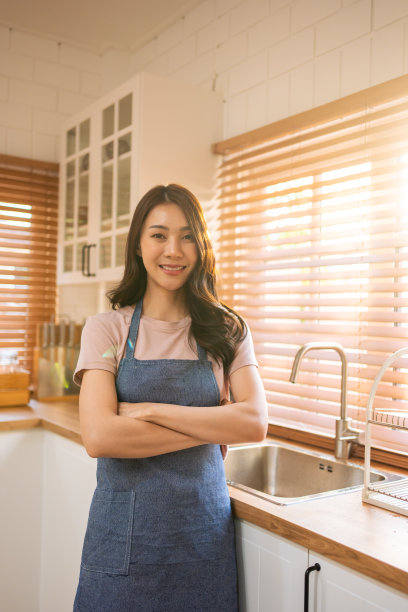 亚洲女性家庭主妇的肖像微笑，看着相机在厨房在家里。漂亮迷人的女管家穿着围裙，自信地挽着胳膊在家里做家务或杂务。图片下载